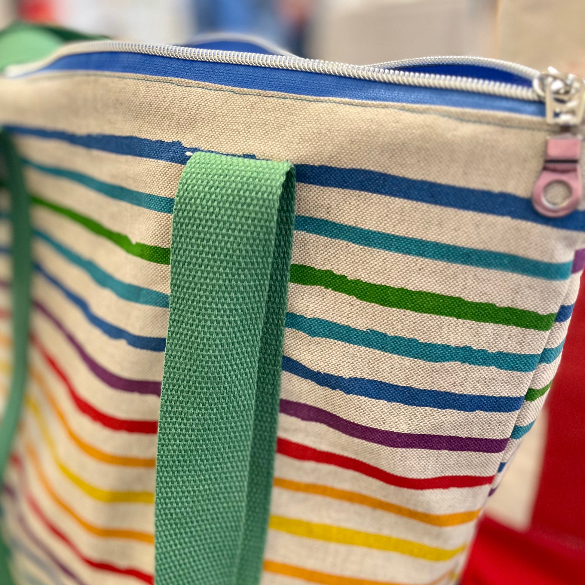 close up of the top zip of a brightly coloured stripey shoulder bag - you can see the silver zipper pull, a blue zip and one of the green shoulder straps hanging down against the rainbow striped bag.