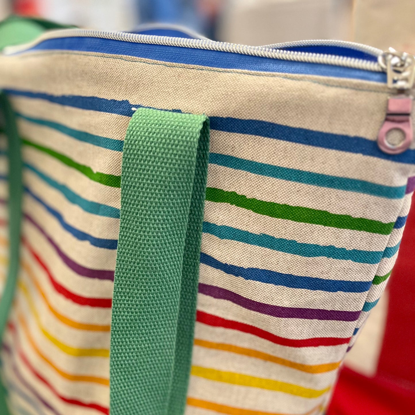 close up of the top zip of a brightly coloured stripey shoulder bag - you can see the silver zipper pull, a blue zip and one of the green shoulder straps hanging down against the rainbow striped bag. 