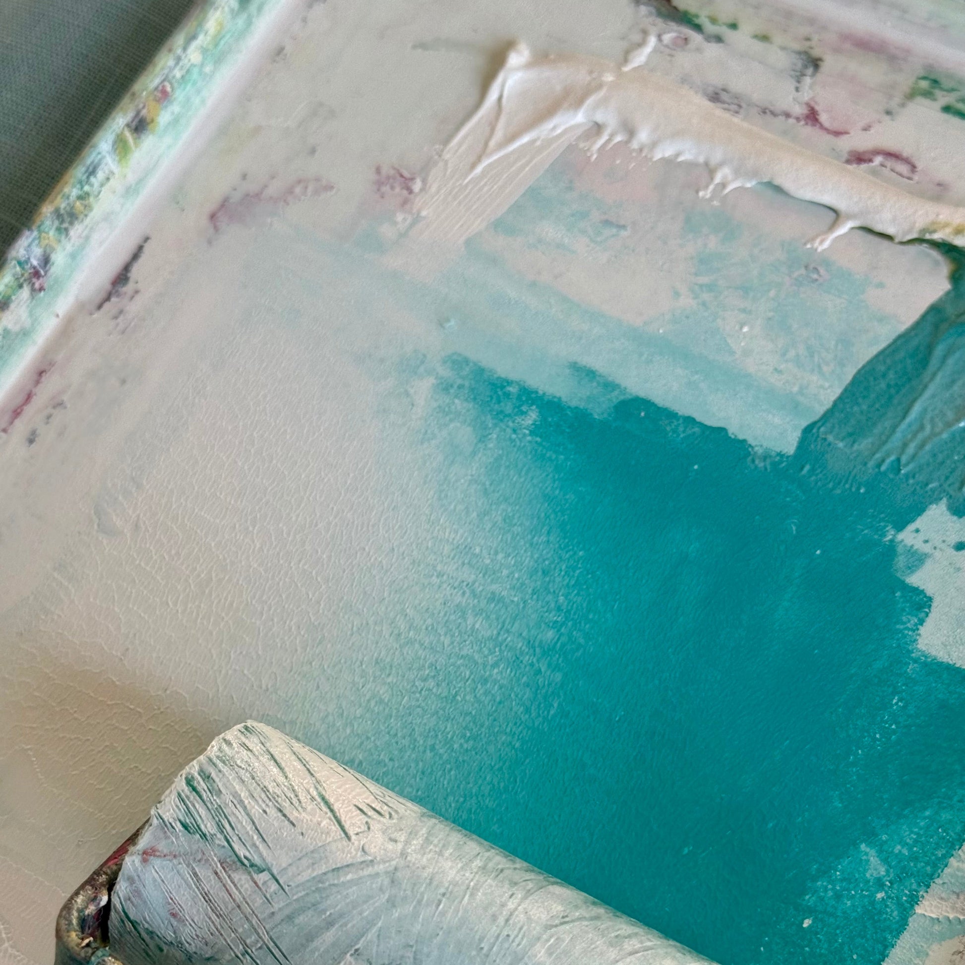 A close-up of a paint roller covered in white paint, resting on a palette with turquoise and off-white paint.
