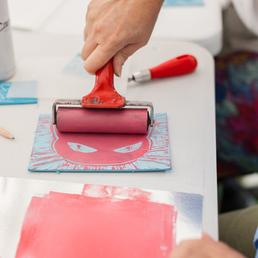 A close-up of a hand holding a red-handled printing roller over a blue and red printed block on a white table, with other crafting tools and materials visible in a real workshop environment.