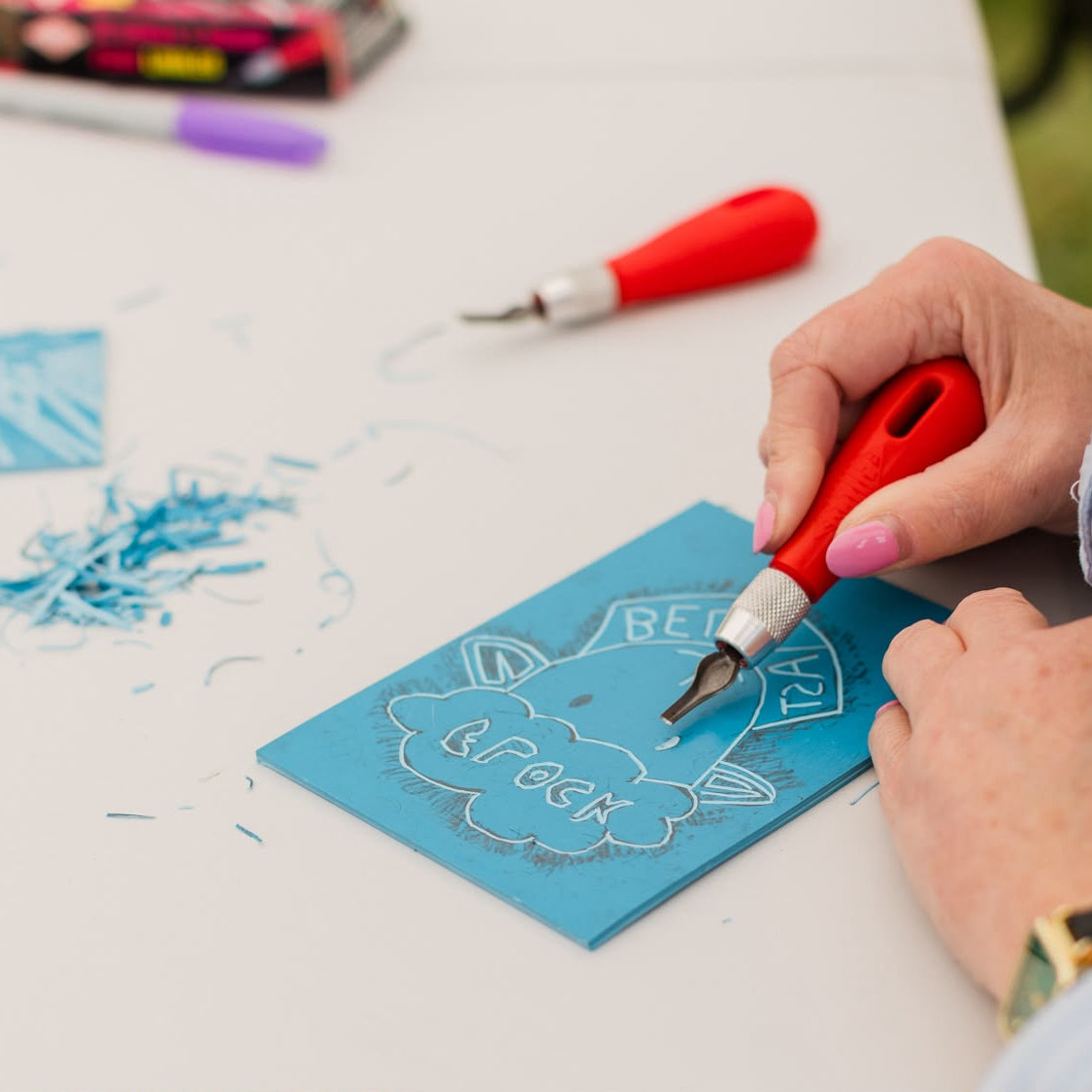 A close-up of a person's hands using a red-handled carving tool to cut a design onto a blue lino sheet on a white table.