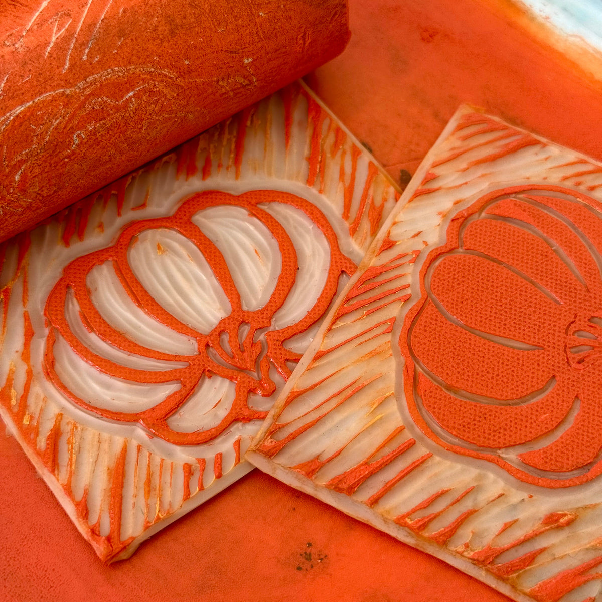 Two carved wooden blocks with pumpkin designs and an orange roller stamp on an orange surface, showing a close-up of the carving and stamping process.