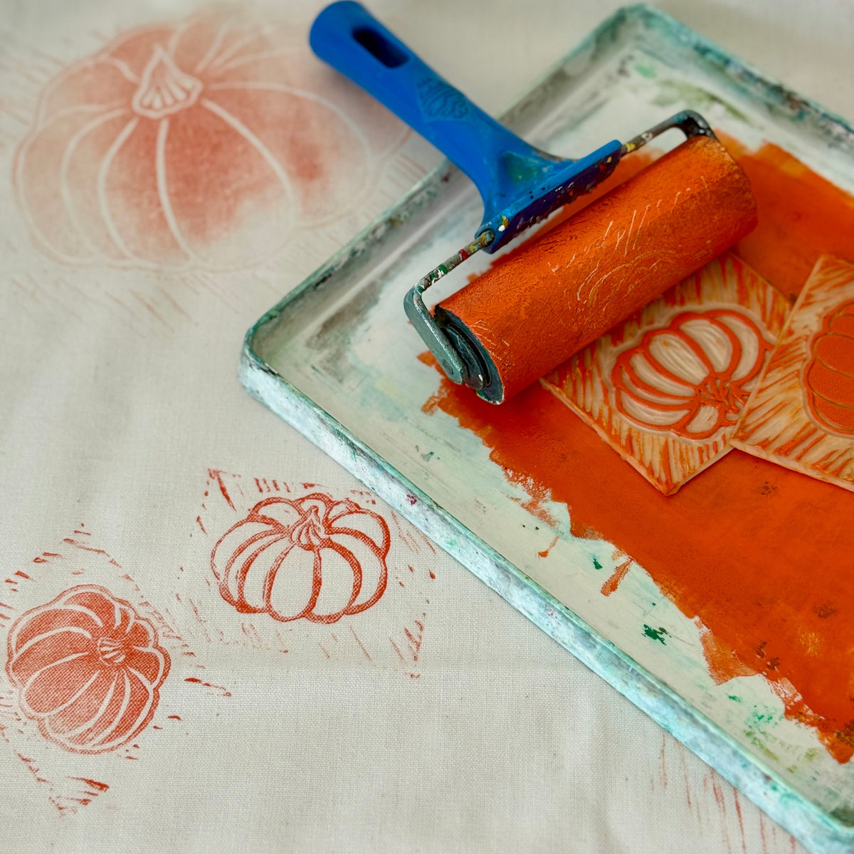 An orange inked roller in a glass tray printing pumpkin motifs onto an off-white fabric surface.