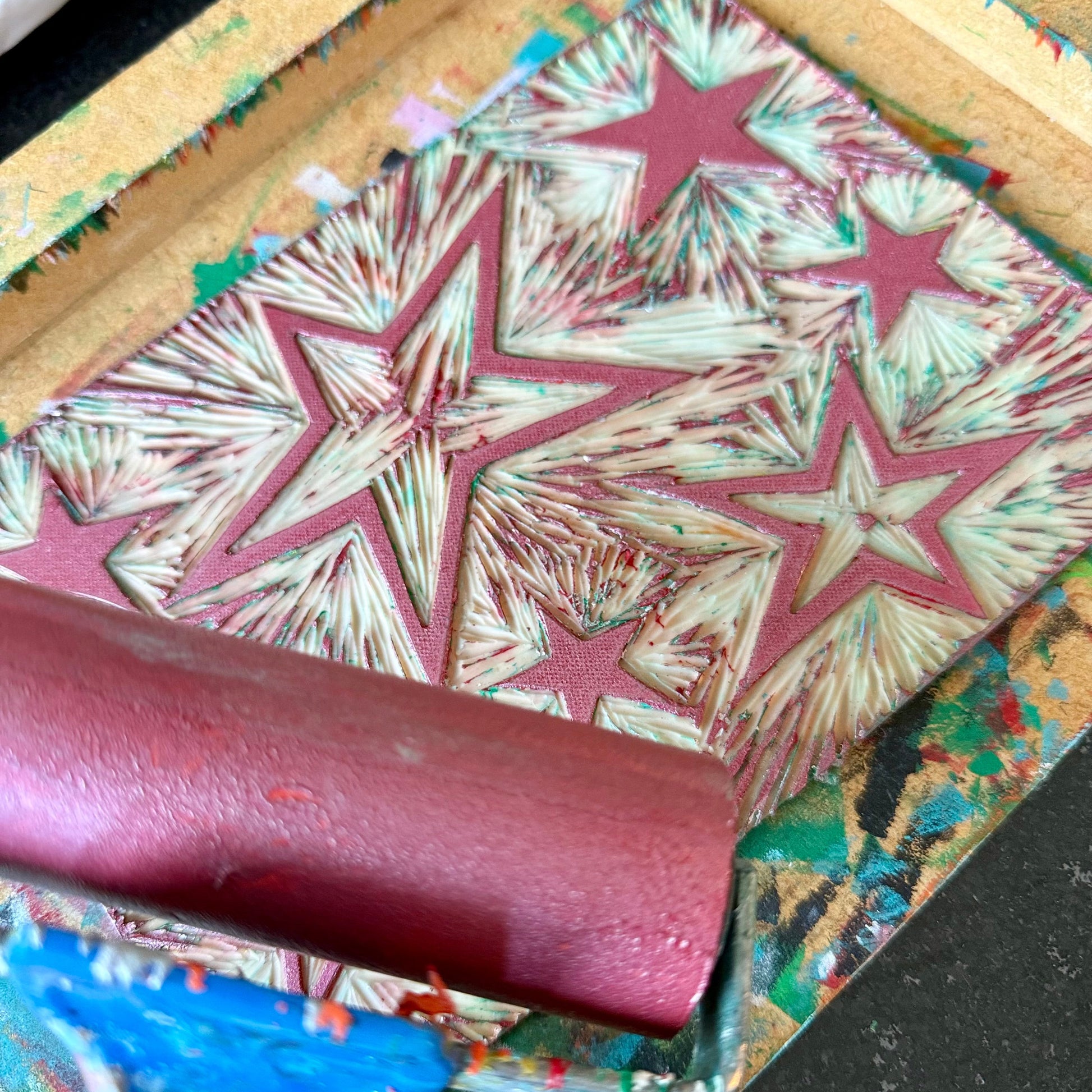 A close-up of a hand-printing or stamping process, showing a wooden frame with a star pattern in pink and cream, being rolled over with a pink roller on a paint-splattered work surface.